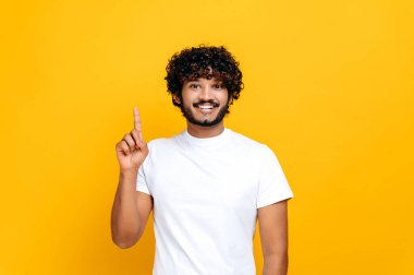 Temel beyaz tişörtlü neşeli Hintli ya da Arap adam, kameraya bakar ve boş fotokopi alanını işaret eder, izole edilmiş turuncu arka planda durur, gülümser. Model-up, boşluk konseptini kopyala