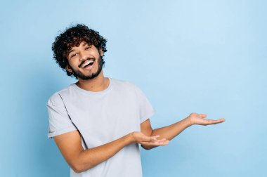 Dost canlısı, kıvırcık saçlı kızılderili ya da Arap tişörtlü adam kameraya bakar ve elleri boş bir yere doğrultur, izole edilmiş mavi arka planda durur, gülümser. Kopyalama alanı kavramı