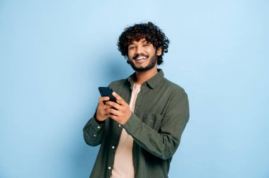 Mutlu yakışıklı, şaşırmış Hintli ya da Arap adam, akıllı telefon kullanıyor, sohbet ediyor, mesaj atıyor, internete, sosyal medyaya bakıyor, izole edilmiş mavi arka planda duruyor, gülümsüyor.
