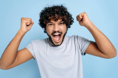 Heyecanlı, şaşırmış Hintli ya da Arap bir adam, basit bir tişörtle, başarı, zafer, kazanma, yumruklarla hareket etme, kameraya bakma, gülümseme, izole edilmiş mavi bir arka planda durma, bağırma