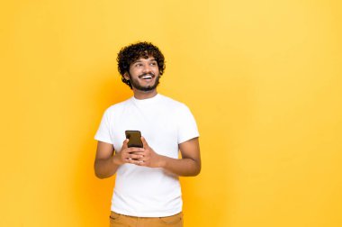 Karizmatik Hintli ya da Arap adam, beyaz tişörtlü, akıllı telefonu elinde tutuyor, internette mesajlaşıyor, internete bakıyor, mutlu mesut yan bakıyor, düşünüyor, turuncu arka planda duruyor, gülümsüyor.