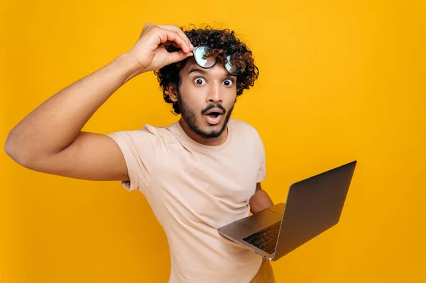 Nadšený šokovaný indický nebo arabský chlap, v ležérním tričku, sundat brýle, drží otevřený notebook v ruce, vypadá překvapeně s otevřenými ústy do kamery, zatímco stojí na izolovaném oranžovém pozadí — Stock fotografie