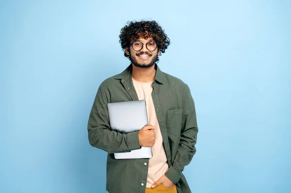 Smart handsome positive indian or arabian guy, with glasses, in casual wear, student or freelancer, holding a laptop in hand, standing on isolated blue background, looking at camera, smiling friendly — Stock Photo, Image