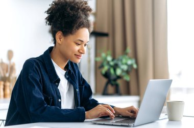 Positive lovely smart african american girl in casual wear, freelancer, student or creative designer, sits at home in the kitchen, uses a laptop, working on a project, browses the Internet, smiles clipart