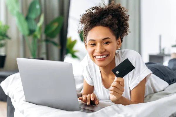 아프리카 계 미국인 행복 한 소녀, 침대에 누워, 손에 신용 카드를 들고, 노트북을 사용하고, 온라인 쇼핑을 하고, 집으로 배달을 주문하고, 무심 한 지불을 요구하고, 카메라를 보고, 웃으면서 — 스톡 사진
