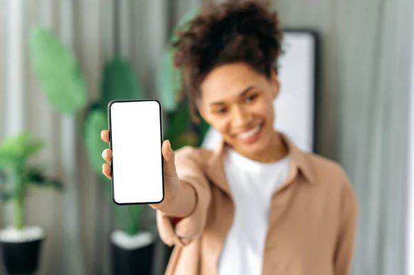 Blurred african american young woman in casual wear, showing modern cell phone with blank white screen, stand at home in a living room, smile, mockup, copy space concept — Stok Foto