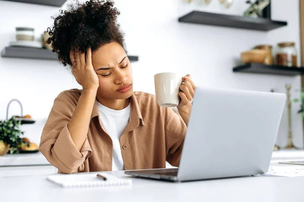 Bored african american girl, freelancer, designer, working remotely from home, tired of boring online work, suffering from chronic fatigue or overwork, holds a cup of coffee in her hand, falls asleep — Stock Photo, Image