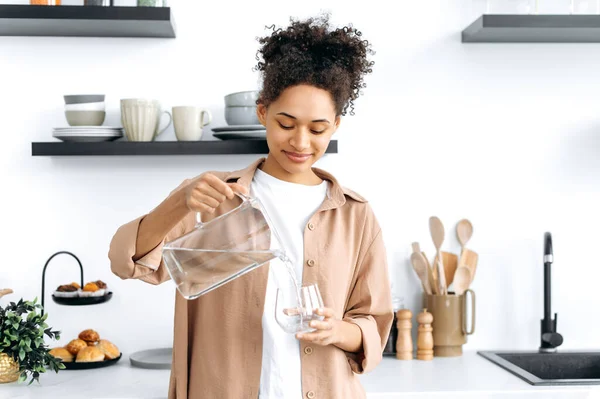 Glückliches gesundes afrikanisch-amerikanisches Mädchen in lässiger Kleidung, zu Hause in der Küche stehend, sauberes Wasser aus einem Krug in ein Glas gießt, täglich viel Wasser trinkt, auf ihre Gesundheit achtet, lächelt — Stockfoto