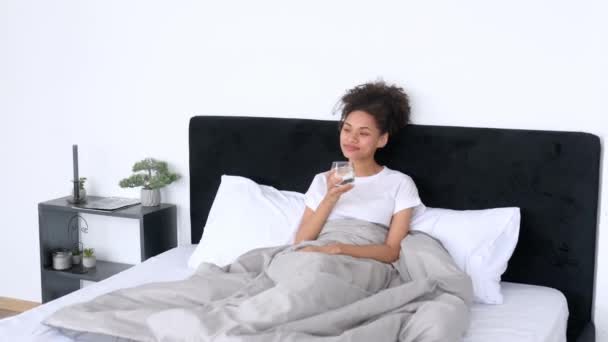 Belle fille afro-américaine satisfaite dans un t-shirt blanc, assise dans un lit le matin, tient un verre d'eau propre, commence la journée avec une norme quotidienne d'eau, tient un mode de vie sain, souriant — Video