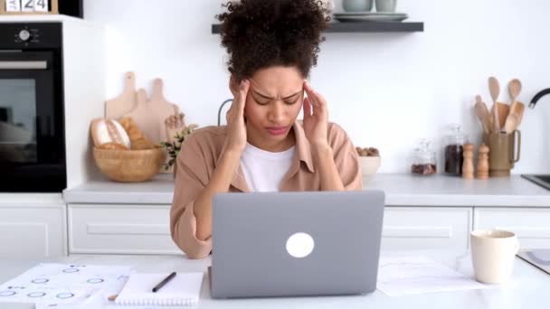 Chica afroamericana cansada trabajando en exceso en una computadora portátil, sentada en una mesa en la cocina en casa, cerró los ojos, masajea sus sienes, tiene migraña, dolor de cabeza, tensión, necesita descanso, descanso — Vídeos de Stock