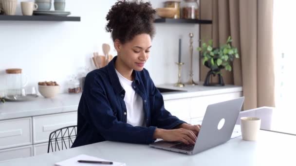 Pozitif, sevimli, Afro-Amerikan kızı, günlük giysiler içinde, serbest çalışan, öğrenci ya da yaratıcı tasarımcı, mutfakta oturuyor, dizüstü bilgisayar kullanıyor, bir proje üzerinde çalışıyor, internette geziniyor, gülümsüyor. — Stok video