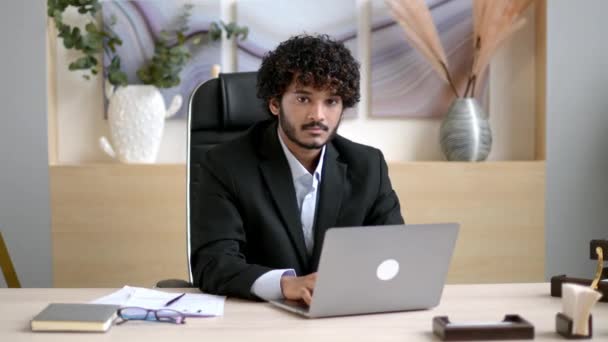 Video portret van jonge volwassen knappe vrolijke zelfverzekerde succesvolle Indiase zakenman, ceo of manager met baard, in zakelijke kleding, zitten aan zijn bureau in het kantoor met een vriendelijke glimlach — Stockvideo