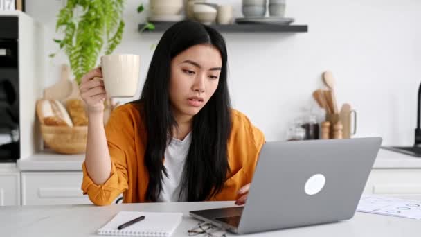 Een slaperig Aziatisch meisje, freelancer of student, zit thuis in de keuken aan een tafel met haar ogen dicht voor een laptop, houdt een kop koffie in haar hand, kan niet wakker worden en aan het werk gaan — Stockvideo