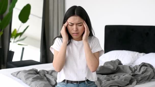 Upprörd frustrerad flicka av asiatisk etnicitet i casual kläder sitter på sängen hemma, har en svår huvudvärk, migrän, masserar hennes tempel, blundar, behöver medicin och vila — Stockvideo