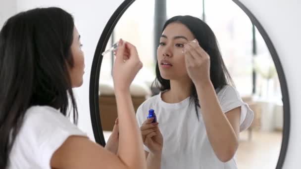 Asian pretty girl in white t-shirt, stands in front of a mirror at home, does her morning beauty routine, takes care of her facial skin, apply moisturizing face serum, prevents wrinkles, smiling — Stock Video