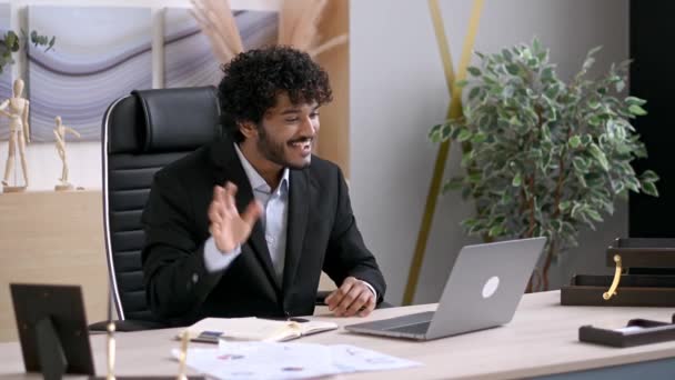 Online videokommunikáció. Pozitív sikeres indiai üzletember hivatalos öltönyben, asztalnál ül az irodában, laptoppal beszél videóhívással, beszél egy kollégájával vagy barátjával, mosolyog. — Stock videók