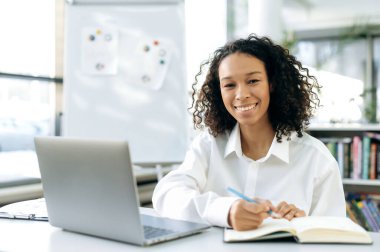 Şirin Afrikalı Amerikalı kadın, Frelancer ya da ofis çalışanı, bir şirkette stajyer, dizüstü bilgisayarında oturuyor, modern resmi giysiler içinde, eğitilmiş, çalışıyor, notlar alıyor, kameraya bakıyor, gülümsüyor.