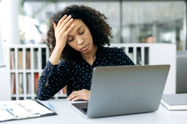 En trøtt, mørkhudet jente, direktør for finansielle konsulenter i et firma, sitter foran en dataskjerm ved et moderne kontor, holder hodet, utslitt av hardt arbeid, trenger hvile – stockfoto