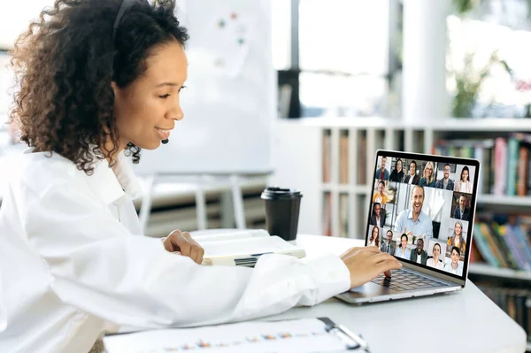Wanita muda african yang sukses, manajer perusahaan, sekretaris, duduk di depan layar laptop, mengadakan konferensi video dengan kelompok kolega internasional, tersenyum ramah — Stok Foto