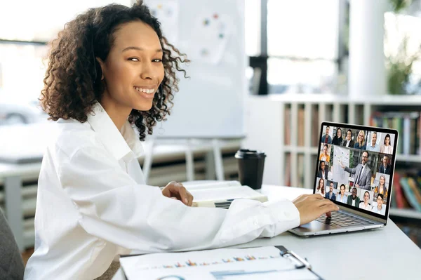Positive successful young adult African American woman, top manager, head of a company, sitting in front of a laptop in a modern office, conducts online briefing with business partners, taking notes — ストック写真