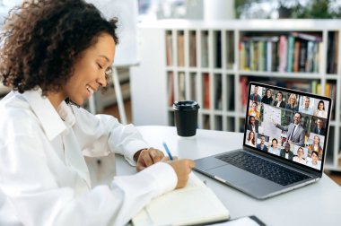 Online eğitim. Zeki Afrikalı Amerikalı kız, öğrenci, yönetici, ekonomist, danışman, bir dizüstü bilgisayarın önünde, modern bir ofiste, internetten eğitim alıyor, dikkatle dinliyor, notlar alıyor.