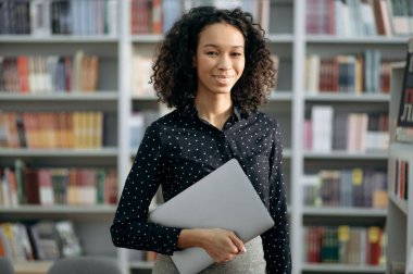 Başarılı zeki Afro-Amerikan genç kadın, öğretmen, öğrenci, modern bir kütüphanede duruyor, iş kıyafetleri giyiyor, elinde dizüstü bilgisayar tutuyor, kameraya bakıyor, gülümsüyor.