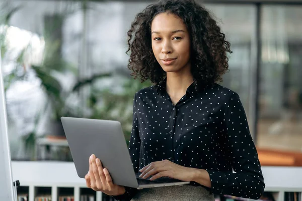 Potret gadis Afrika Amerika yang sukses, auditor, freelance, pemasaran, manajer, berdiri di kantor modern mengenakan pakaian bisnis bergaya, memegang laptop di tangannya, tersenyum menyenangkan — Stok Foto
