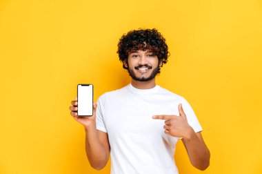 Şaşırmış Hintli ya da Arap kıvırcık tişörtlü adam, kameraya bakıyor, akıllı telefonu boş beyaz ekran ile tutuyor ve işaret ediyor, izole edilmiş turuncu arka planda duruyor, gülümsüyor.