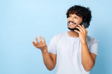 Cep telefonu sohbet konsepti. Mutlu Arap ya da Hintli çekici adam, akıllı telefonunu kullanıyor, akıllı telefonuyla konuşuyor, izole edilmiş mavi arka planda duruyor, başka tarafa bakıyor, gülümsüyor dostane bir şekilde el kol işareti yapıyor.