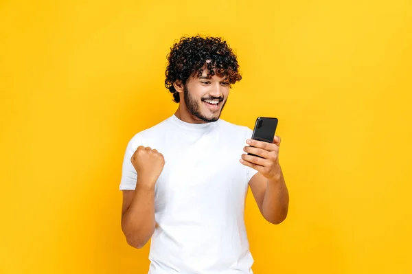 Šťastný radostně vzrušený indický nebo arabský chlap drží smartphone, získat nečekané zprávy, výhra loterie, stojí na izolovaném oranžovém pozadí, veselý výraz obličeje, zubatý úsměv, gestikulace s pěstí — Stock fotografie