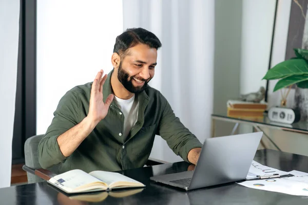 Agradável cara índio amigável, gerente ou consultor financeiro, se comunica com os colegas em uma chamada de vídeo, discute resultados e estratégia em um escritório moderno, negociações remotas, gesto de saudação — Fotografia de Stock