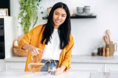 Günlük su girişi. Pozitif, şık giysiler içinde güzel bir Çinli kız, evde mutfakta duruyor, bir sürahiden bardağa su dolduruyor, kameraya bakıyor, gülümsüyor. Sağlıklı yaşam tarzı