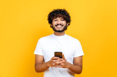 Yakışıklı Hintli ya da Arap adam akıllı telefonunu kullanıyor, arkadaşlarıyla mesajlaşıyor, internette ve sosyal ağlarda geziniyor, izole edilmiş turuncu arka planda duruyor, kameraya bakıyor, gülümsüyor.