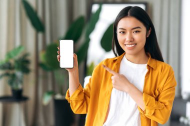 Güzel, mutlu bir Çinli kız, günlük giysiler içinde, evinde duruyor, elinde akıllı telefonuyla elinde boş bir beyaz model ekran ile, kameraya bakıyor, gülümsüyor.