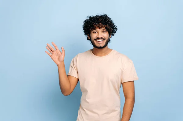 Handsome positive arabian or indian curly-haired guy, wearing casual t-shirt, standing on isolated blue background, looking at the camera, waving hand, welcome sign, smiling friendly — Stock Photo, Image