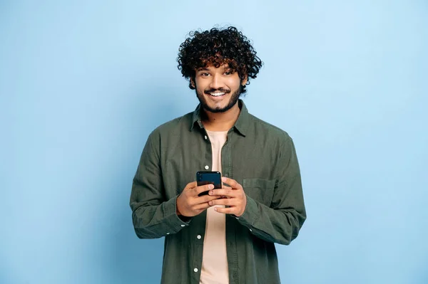 Handsome positive millennial indian or arabian guy casual wear, uses smartphone, messaging with friends online, browses social networks, news, looks at camera, smile, stand on isolated blue background — Stock Photo, Image