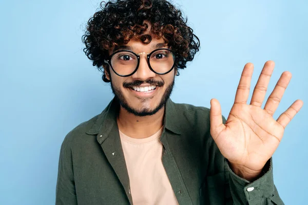 Close-up de carismático atraente positivo encaracolado indiano ou árabe cara na camisa casual com óculos, olhando para a câmera, sorrindo alegremente, acenando, gesto de saudação, ficar em um fundo azul isolado — Fotografia de Stock