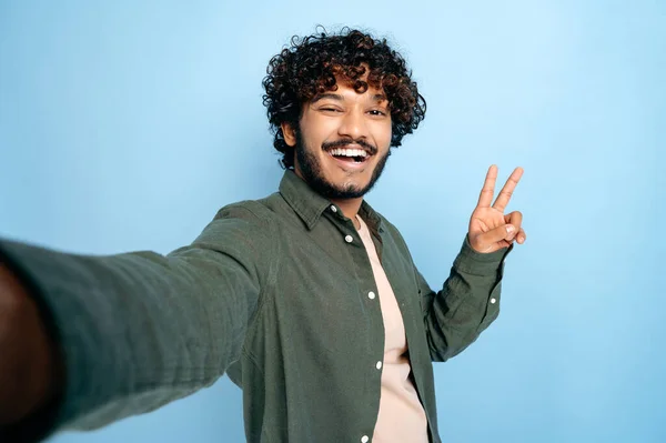 Cheerful happy indian or arabian excited guy with gladden face expression, in casual clothes, doing selfie shot on smartphone and smiles into the camera, shows peace symbol, isolated blue background — Stock Photo, Image