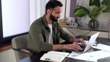 Konsantre olmuş zeki Hintli ya da Arap işadamı, tüccar, simsar, modern bir ofiste bir iş yerinde oturuyor, istatistiklere odaklanıyor, finansal tablolara bakıyor, bir dizüstü bilgisayarda çalışıyor.