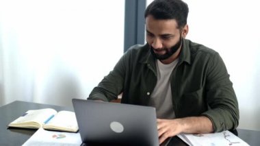 Pozitif zeki Hintli ya da Arap, günlük kıyafetler içinde, serbest çalışan ya da yönetici, ofiste çalışma masasında oturmuş, webinar dinliyor, notlar alıyor, öğreniyor, becerilerini geliştiriyor, gülümsüyor.