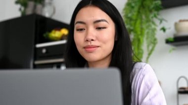 Güzel, pozitif Asyalı esmer kız, tasarımcı, serbest çalışan ya da öğrenci, mutfakta dizüstü bilgisayarla oturuyor, çalışıyor ya da uzaktan çalışıyor, dizüstü bilgisayar ve internet kullanıyor, hayal kuruyor, düşünüyor, gülümsüyor.