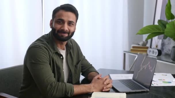 Portret odnoszącego sukcesy indyjskiego lub arabskiego przedsiębiorcy, maklera giełdowego, inwestora, siedzącego przy biurku w biurze, patrzącego w kamerę, uśmiechniętego przyjaznego. Wykresy kryptograficzne na ekranie laptopa — Wideo stockowe