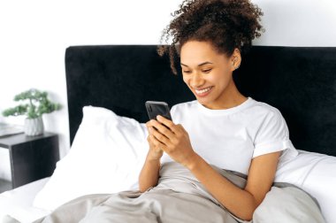 Akıllı telefon kullanıyor. Mutlu, Afro-Amerikan genç bir kadın cep telefonunu evde yatakta yatarken kullanıyor cep telefonuyla mesajlaşıyor, arkadaşlarıyla sohbet ediyor, sosyal medyada geziniyor, gülümsüyor.