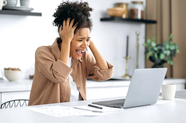 Frustrasi kecewa Afrika gadis berambut cokelat, freelancer, manajer, duduk di meja di dapur, menggunakan laptop sementara pekerjaan jauh, terkejut oleh berita atau pesan, menderita kerugian keuangan, berteriak — Stok Foto