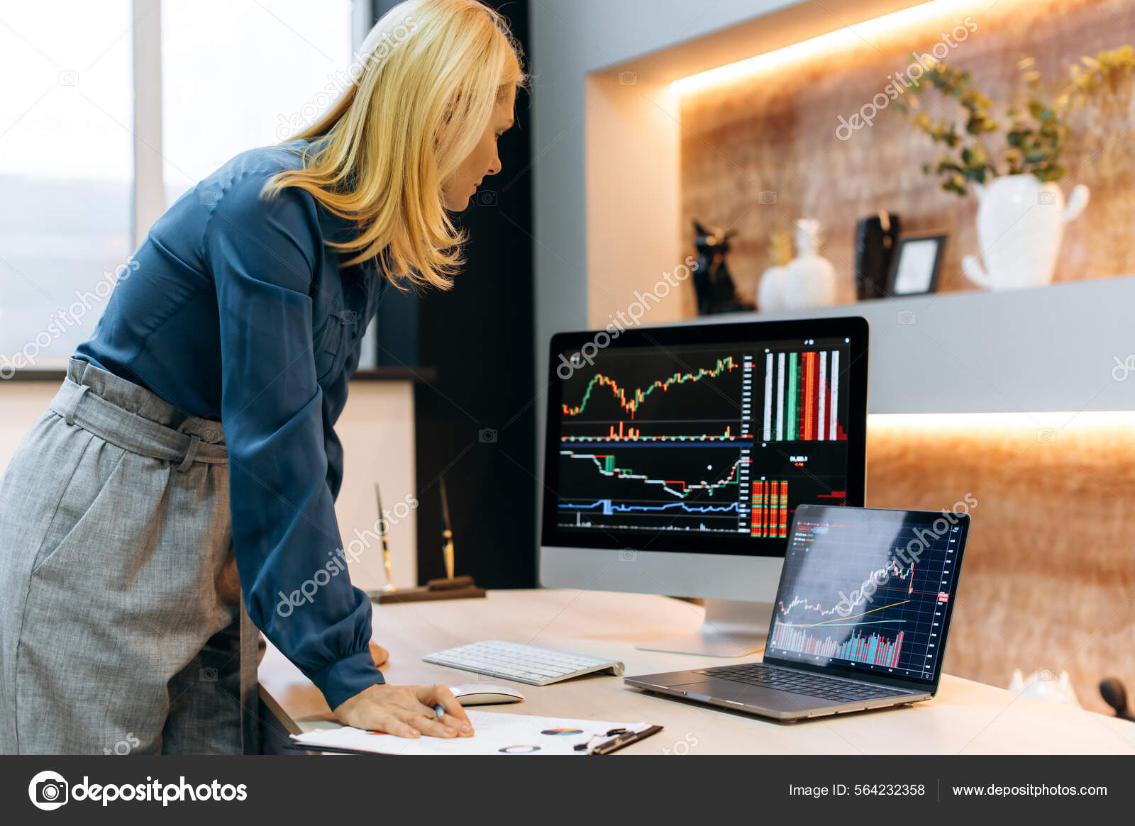 成熟的高加索聪明、成功的金发女人，股票交易员、密码投资者，站在台式机旁边，在电脑和笔记本电脑屏幕上分析图表，比较指标，评估风险——
