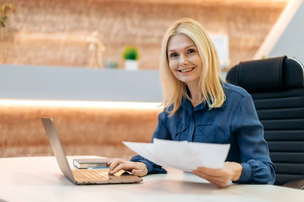 Portret van intelligente elegante volwassen blanke positieve vrouwelijke werknemer, zakenvrouw, bankier, zit aan het bureau in het kantoor, werken met documenten en laptop, kijkt naar de camera, glimlachend vriendelijk — Stockfoto