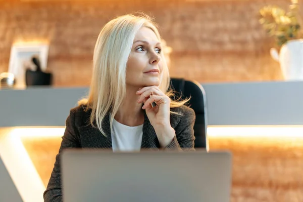 Mujer rubia de mediana edad pensativa, director general, gerente, corredor, banquero se sienta en una mesa en la oficina moderna, vestido con traje formal, piensa en una idea de negocio o estrategia financiera, planificación, mira a un lado —  Fotos de Stock