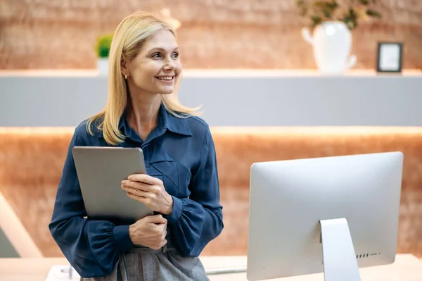 Elegante succesvolle intelligente volwassen blonde blanke zakenvrouw, top manager, bankier, makelaar, staat in modern kantoor, in een blauw shirt, laptop vasthoudend, naar de zijkant kijkend, lachend vriendelijk — Stockfoto