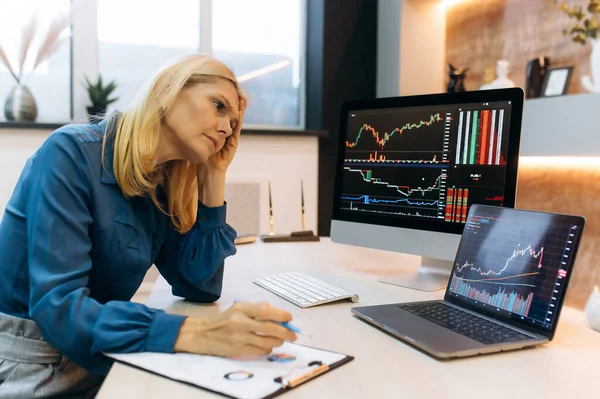 Tired overworked sad mature caucasian smart blonde woman, a stock trader, a crypto investor, sit a the desktop, analyzes charts on a computer and laptop screen, compares indicators, assesses risks — Stock Photo, Image