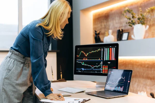 Rijpe blanke slimme succesvolle blonde vrouw, een aandelenhandelaar, een crypto investeerder, staat in de buurt van het bureaublad, analyseert grafieken op een computer en laptop scherm, vergelijkt indicatoren, beoordeelt risico 's — Stockfoto